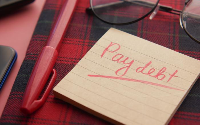 a note that says pay debt next to a pen and glasses by Towfiqu barbhuiya courtesy of Unsplash.